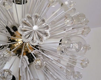 May include: A white and gold chandelier with clear glass crystals and light bulbs. The chandelier has a starburst design with many arms extending outward.