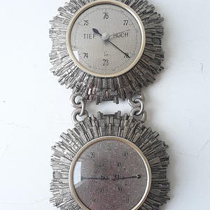 May include: A silver-toned barometer with a chain hanging from the top. The barometer has two circular faces, one with a needle pointing to "76" and the other with a needle pointing to "60". The face with the needle pointing to "76" has the words "TIEF" and "HOCH" printed on it. The face with the needle pointing to "60" has numbers printed around the edge.