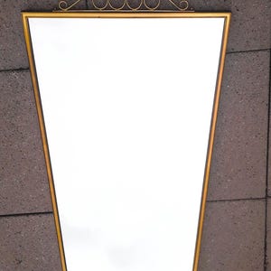May include: A gold-framed mirror with a unique trapezoid shape. The mirror has a decorative gold scrollwork design at the top. The mirror is a decorative home decor item.