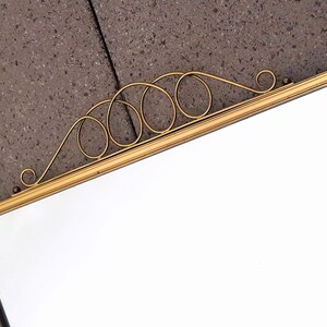 May include: A rectangular mirror with a gold-colored frame and decorative scrollwork at the top. The mirror's surface is clear and reflective, set against a textured gray background. The frame has a simple, elegant design.