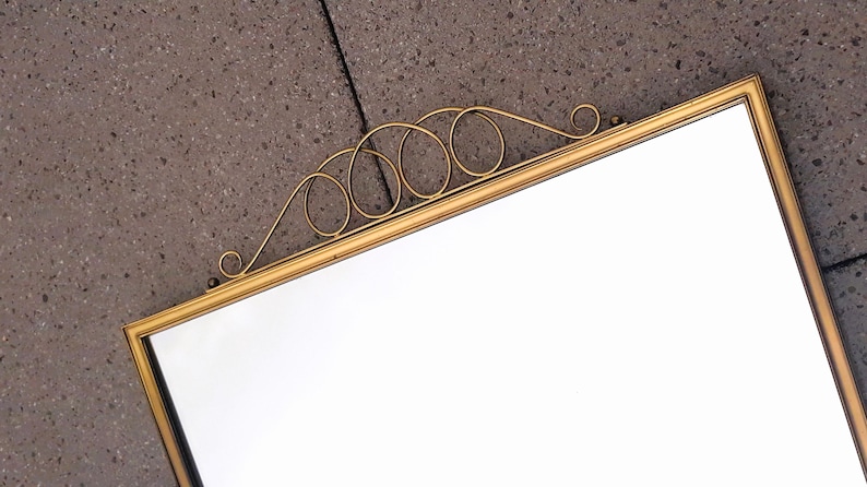 May include: A rectangular mirror with a gold-colored frame and decorative scrollwork at the top. The mirror's surface is clear and reflective, set against a textured gray background. The frame has a simple, elegant design.