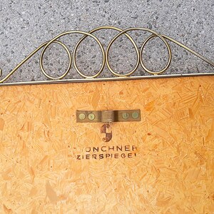 May include: A vintage decorative mirror with a gold-colored metal frame. The frame features a series of interconnected oval shapes and a decorative flourish. The wooden backing has a metal bracket and the text "UNCHNER ZIERSPIEGEL".