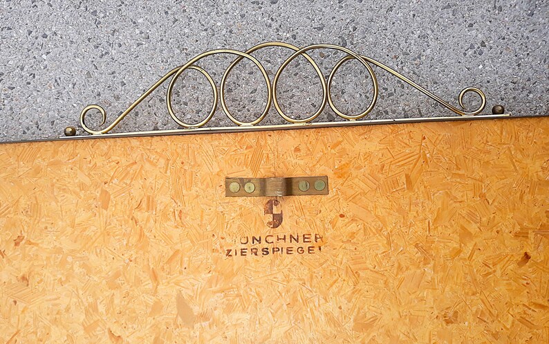 May include: A vintage decorative mirror with a gold-colored metal frame. The frame features a series of interconnected oval shapes and a decorative flourish. The wooden backing has a metal bracket and the text "UNCHNER ZIERSPIEGEL".