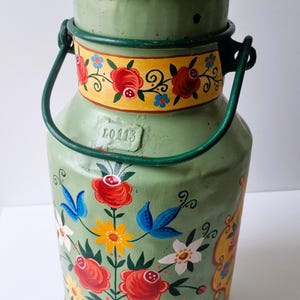 Op de afbeelding: Een saliegroene metalen melkbus met een handgeschilderd bloemmotief in rood, geel en blauw. De bus heeft een afneembaar deksel met handvat, een zwenkbare beugel en de in reliëf aangebrachte tekst LO113 op de voorzijde. De onderkant is donkergroen.