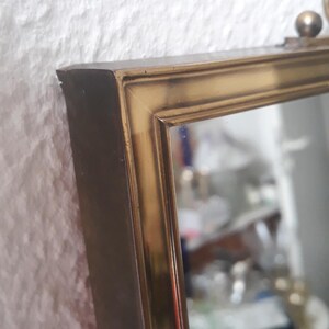 May include: A rectangular mirror with a gold-colored frame. The frame has a beveled edge and a small decorative ball at the top. The mirror is set against a textured white wall.