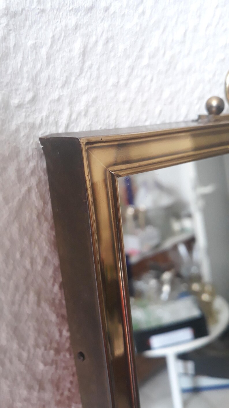 May include: A rectangular mirror with a gold-colored frame. The frame has a beveled edge and a small decorative ball at the top. The mirror is set against a textured white wall.