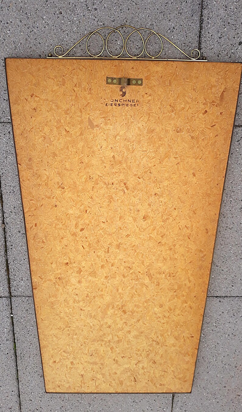May include: Back of a vintage mirror with a trapezoid shape. The mirror's backing is made of light brown particle board. A decorative gold-colored metal piece with a series of circles is at the top. The text "MUNCHNER ZIERSPIEGEL" is visible.