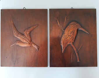 May include: Two wooden wall plaques with raised copper relief sculptures of birds. The plaque on the left features a bird in flight, while the plaque on the right features a bird perched on a branch.