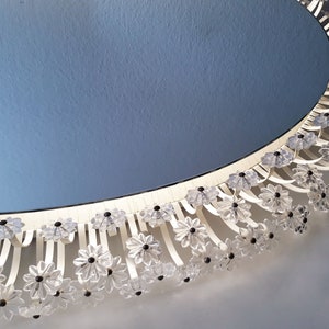 Peut inclure: Un miroir ovale avec un cadre blanc et une bordure florale décorative. La bordure présente des cristaux transparents en forme de fleurs avec des centres noirs. Le miroir est posé contre un mur bleu clair.