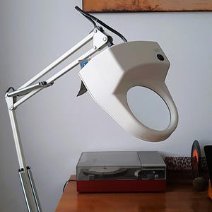 Puede incluir: Una lámpara de aumento blanca con un brazo ajustable, colocada sobre un escritorio de madera. Un tocadiscos con base roja está sobre el escritorio. Un mapa del mundo está en la pared de fondo.