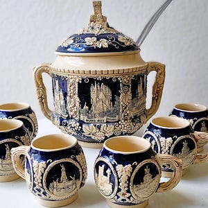 May include: A vintage ceramic soup tureen with lid and six matching mugs. The set features a cream and navy blue color scheme with relief designs of buildings, grapes, and the text "BURG ELTZ MOSSEL".