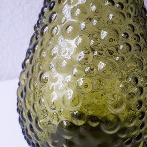May include: A green glass bottle with a textured surface. The bottle has a round, pear-shaped body with a wide base and a narrow neck.