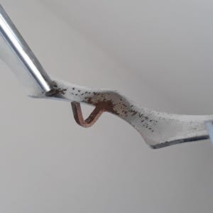 May include: Close-up of a silver metal hook with a rusty brown finish. The hook is bent at a 90-degree angle and has a small loop at the end.