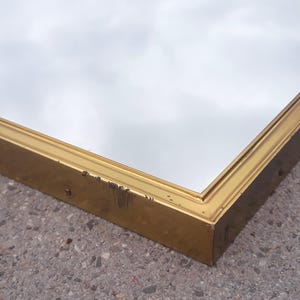 May include: A rectangular mirror with a gold-colored frame. The frame has some wear and tear. The mirror reflects a cloudy sky. The mirror is placed on a gray, textured surface.