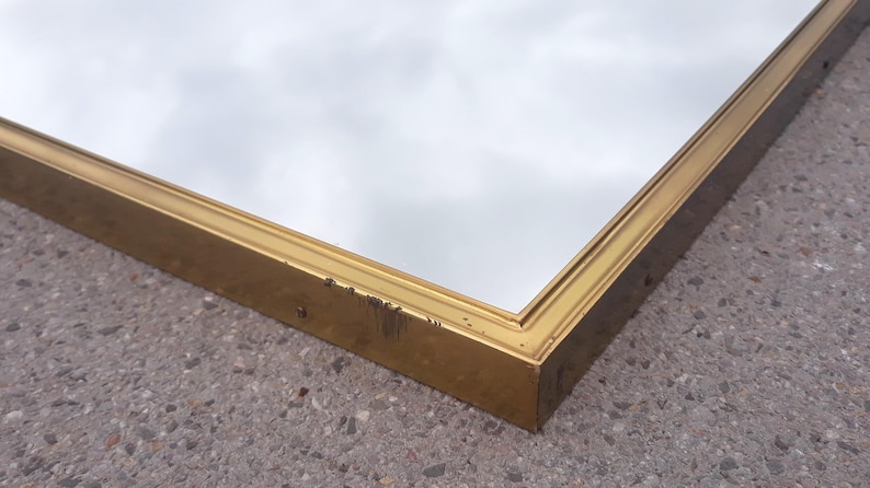 May include: A rectangular mirror with a gold-colored frame. The frame has some wear and tear. The mirror reflects a cloudy sky. The mirror is placed on a gray, textured surface.