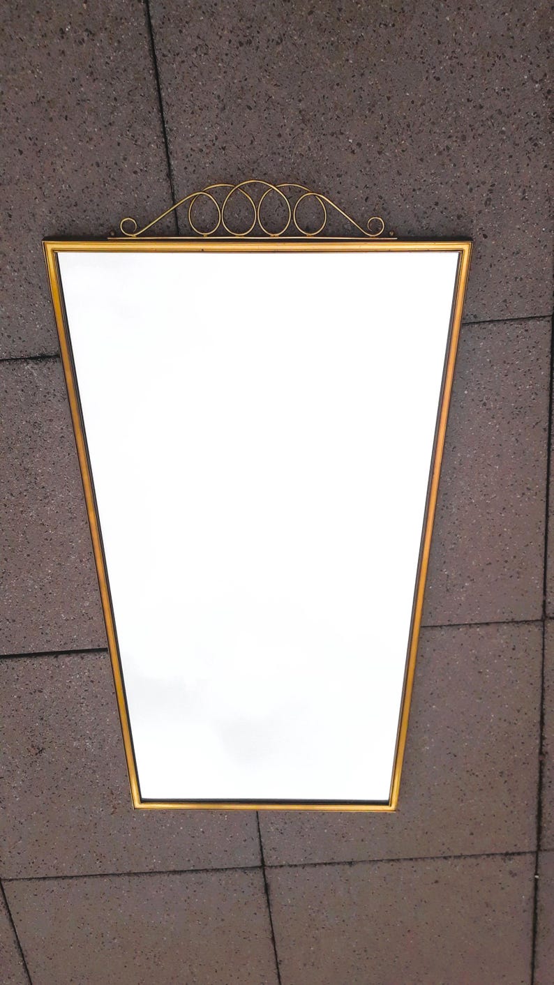 May include: A gold-framed mirror with a unique trapezoid shape. The mirror has a decorative gold scrollwork design at the top. The mirror is a decorative home decor item.