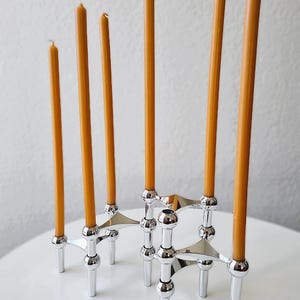 May include: A silver-colored, geometric candle holder with six slender, orange candles. The holder has a modern design with multiple arms and a polished finish. The candles are evenly spaced, creating a symmetrical display.