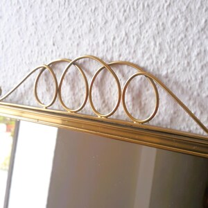 May include: A rectangular mirror with a gold-colored frame and a decorative gold metal top. The top features a series of interconnected oval shapes and a curved design. The mirror is set against a white textured wall.