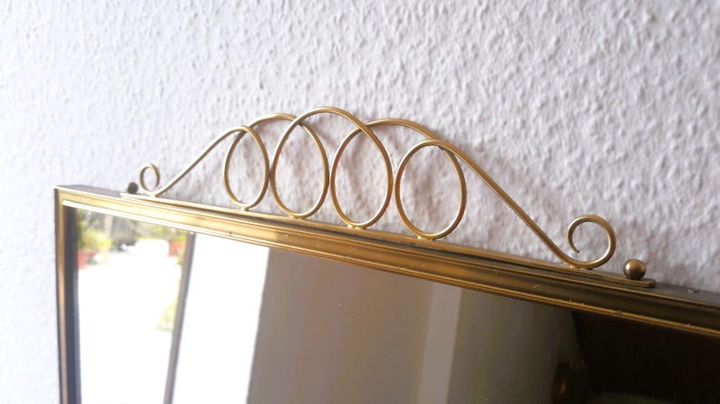 May include: A rectangular mirror with a gold-colored frame and a decorative gold metal top. The top features a series of interconnected oval shapes and a curved design. The mirror is set against a white textured wall.