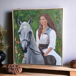 Puede incluir: Pintura al óleo de una mujer montando un caballo blanco. La mujer tiene el pelo largo y ondulado de color castaño rojizo y lleva un vestido blanco con un chaleco azul. El caballo está frente a un fondo verde con flores. La pintura está en un marco dorado.