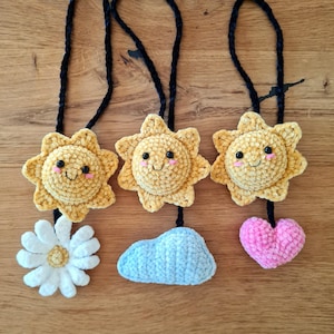 Puede incluir: Tres amuletos de sol tejidos a crochet con caras sonrientes, cada uno unido a un cordón negro. Los amuletos de sol son amarillos y tienen mejillas rosadas. Los cordones cuelgan de una superficie de madera. Debajo de los amuletos de sol hay tres amuletos tejidos a crochet: una margarita blanca, una nube azul y un corazón rosa.