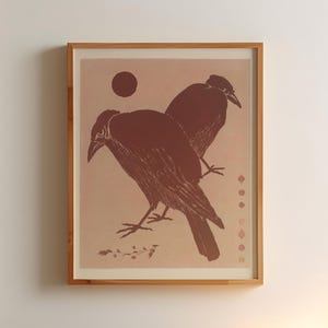May include: A framed print of two crows in a minimalist style. The crows are depicted in a dark red color against a light pink background. The print is framed in a natural wood frame.