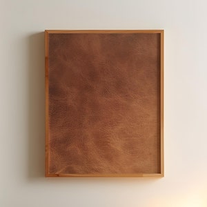 Könnte beinhalten: Ein braunes, ledergerahmtes Bild hängt an einer weißen Wand über einem hölzernen Nachttisch mit einer Lampe und einem weißen Bett mit einer braunen Decke.