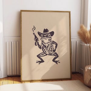 Può includere: Stampa incorniciata con un'illustrazione stilizzata di una rana con cappello da cowboy e pistola fumante. La rana è blu scuro su sfondo beige, con cornice in legno. L'opera d'arte ha un'estetica rustica e vintage.