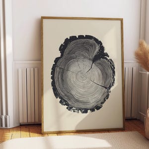 May include: A framed print of a tree trunk cross-section in black and white. The artwork displays detailed growth rings and a cracked centre, set against a cream background. The frame is light brown. The print is leaning against a white wall.