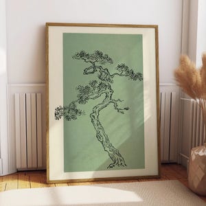 Könnte beinhalten: Ein gerahmter Kunstdruck mit einer schwarzen Tuschezeichnung eines knorrigen, windschiefen Baumes auf einem salbeigrünen rechteckigen Hintergrund. Das Bild befindet sich in einem hellen Holzrahmen mit einem breiten, cremeweißen Passepartout.
