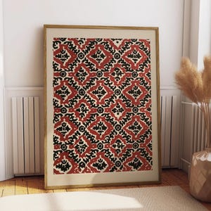 May include: A framed print featuring a repeating pattern of red, black, and cream-colored shapes. The design includes diamond and floral motifs. The artwork is displayed in a light-colored wooden frame, leaning against a white wall and a radiator.