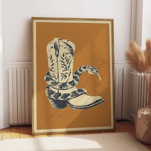 May include: Framed art print featuring a cream-colored cowboy boot with dark blue floral detailing, entwined by a dark blue and cream snake. The artwork is set against an orange background, with a wooden frame.