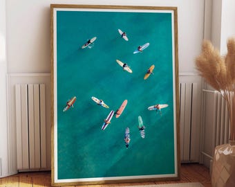 May include: A framed print featuring an aerial view of surfers on surfboards in turquoise water. The surfboards are various colors and designs, with people on them. The frame is a light brown color.