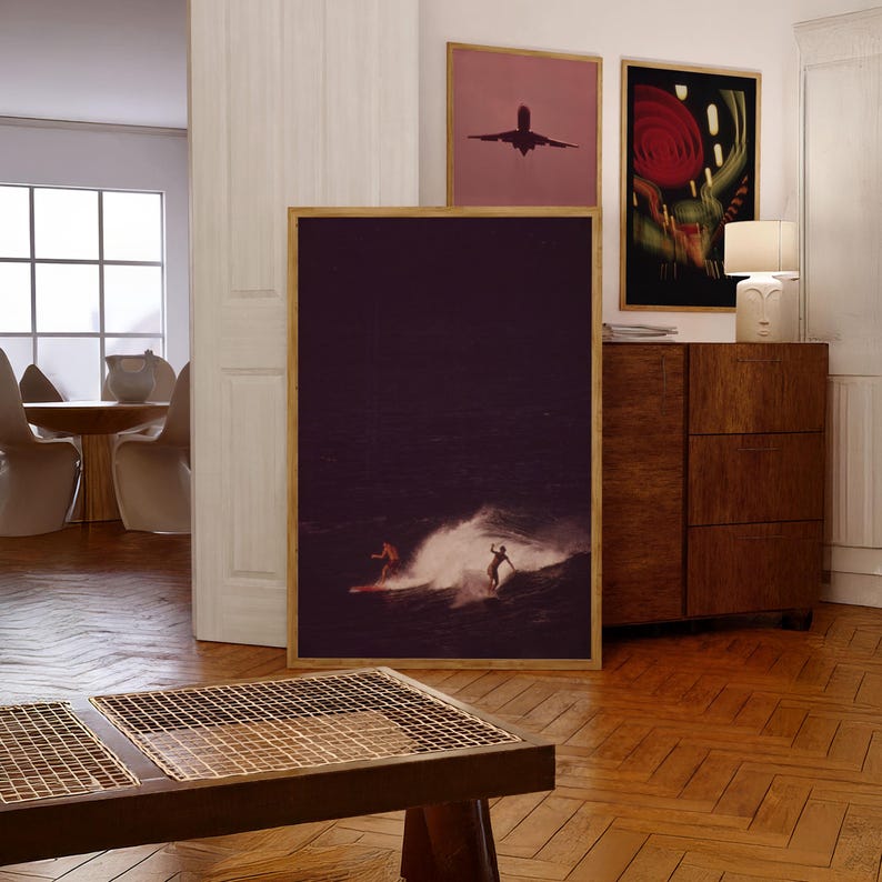 May include: A framed photograph of two people surfing in a dark ocean with a wave breaking behind them. The photograph is hanging on a white wall in a living room with a wooden cabinet and a lamp.