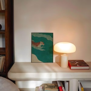 May include: A framed art print featuring a tiger swimming in a teal-colored body of water. The artwork is displayed on a white shelf, next to a white lamp with a rounded shade and a red book.