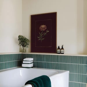 May include: A bathroom scene featuring a white bathtub with a dark green towel, a stack of white towels, and a framed botanical print. The wall has green and white vertical tile and a shelf with two brown bottles and a small plant.