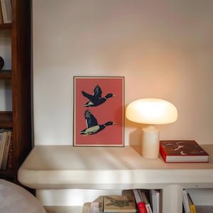 May include: A framed art print featuring two ducks in flight against a red background. A white mushroom-shaped lamp and a red book are on a white shelf. Books are visible on the shelf below.