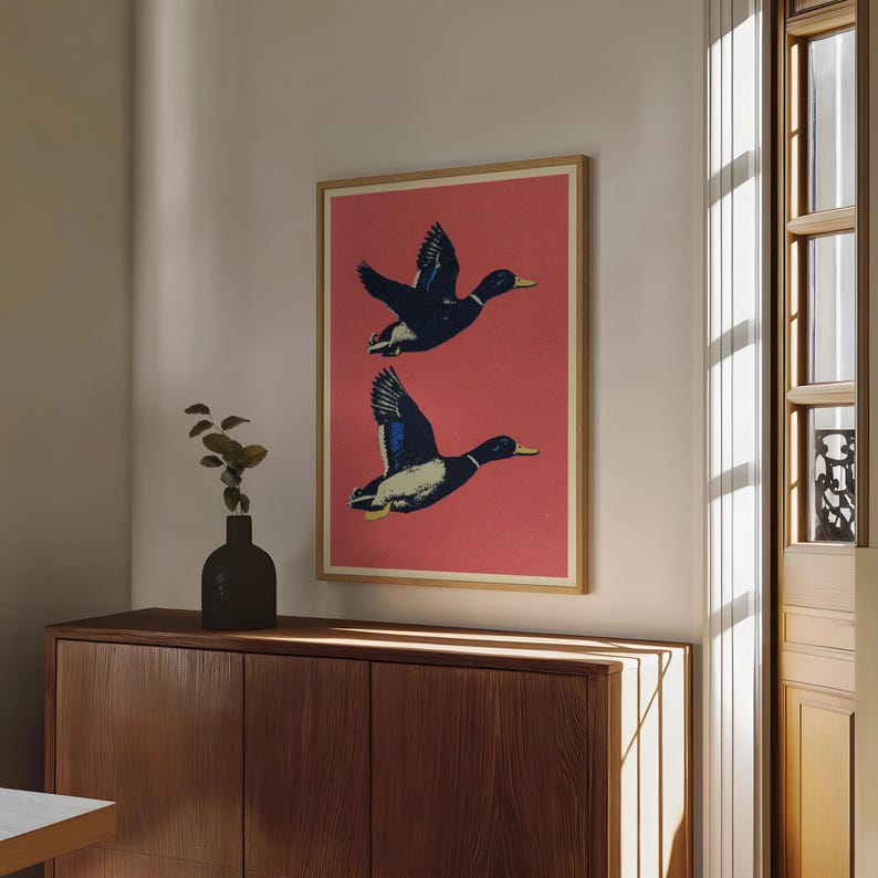 May include: A framed art print featuring two ducks in flight against a red background. The ducks are depicted in shades of black, blue, and white, with yellow beaks. The artwork is displayed in a room with a wooden cabinet and a window.