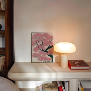 May include: A framed artwork depicts a pink flowering tree with a black frame. A white mushroom-shaped lamp is lit, next to a red book. The scene is set on a white shelf with books below, creating a cozy, artistic ambiance.