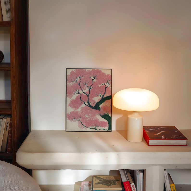 May include: A framed artwork depicts a pink flowering tree with a black frame. A white mushroom-shaped lamp is lit, next to a red book. The scene is set on a white shelf with books below, creating a cozy, artistic ambiance.