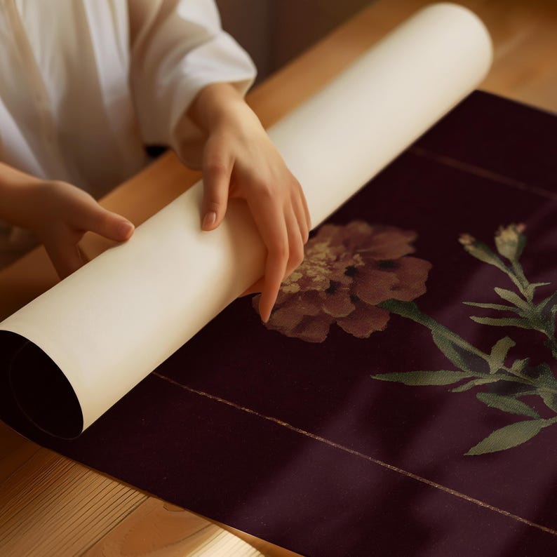 May include: A person is unrolling a cream-colored paper scroll onto a dark burgundy surface with a floral print. The floral print features a large, detailed flower in shades of burgundy and brown, with green leaves and stems.