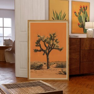 May include: A framed print of a Joshua Tree in a desert landscape. The background is a vibrant orange color. The tree is green and has a thick trunk. The print is in a wooden frame.