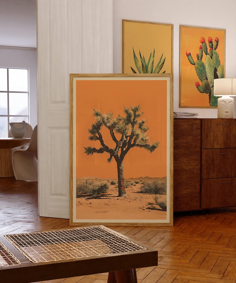 May include: A framed print of a Joshua Tree in a desert landscape. The background is a vibrant orange color. The tree is green and has a thick trunk. The print is in a wooden frame.