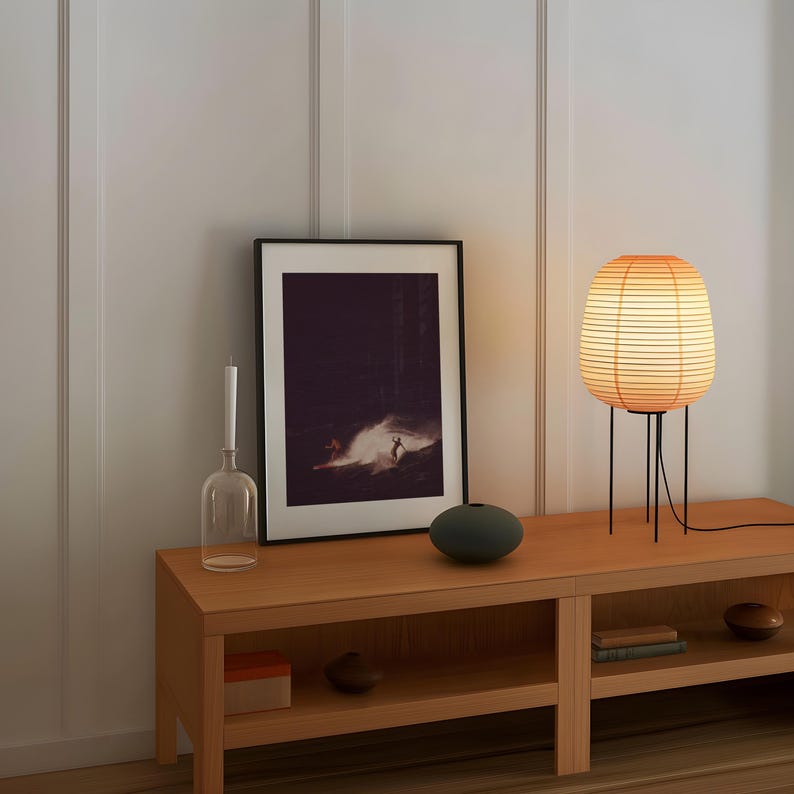 May include: A wooden shelf unit with two shelves, a black framed picture of a surfer, a clear glass bottle, a dark gray round vase, and a table lamp with a light brown paper shade.