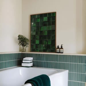 May include: A bathroom scene featuring a white bathtub with a dark green towel, and a wall-mounted artwork composed of green square tiles within a wooden frame. The wall has green and white vertical tiles. Two brown bottles sit on a shelf.
