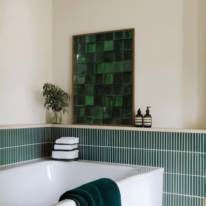May include: A bathroom scene featuring a white bathtub with a dark green towel, and a wall-mounted artwork composed of green square tiles within a wooden frame. The wall has green and white vertical tiles. Two brown bottles sit on a shelf.