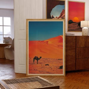 K&ouml;nnte beinhalten: Ein gerahmter Druck einer W&uuml;stenlandschaft mit einem Kamel, das &uuml;ber die Sandd&uuml;nen geht. Der Himmel ist blau und der Sand ist orange.