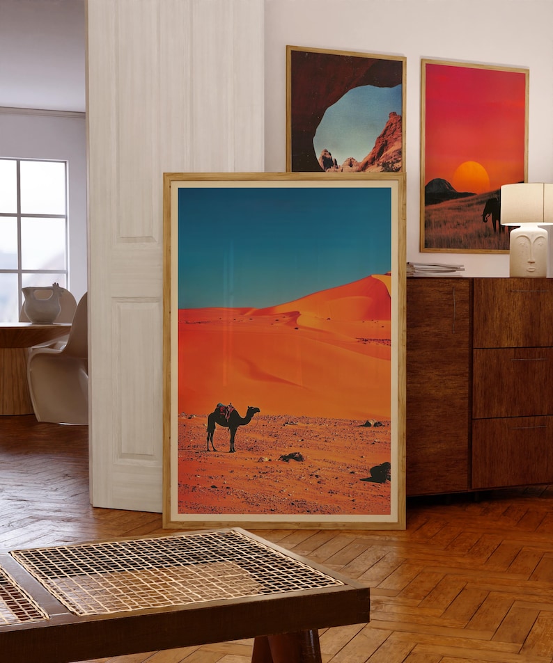 K&ouml;nnte beinhalten: Ein gerahmter Druck einer W&uuml;stenlandschaft mit einem Kamel, das &uuml;ber die Sandd&uuml;nen geht. Der Himmel ist blau und der Sand ist orange.