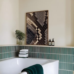Op de afbeelding: Een rechthoekig wandkunstwerk met een gedetailleerd slangenleerpatroon in bruin-, grijs- en cr&egrave;metinten, gevat in een dunne houten lijst. Het kunstwerk hangt boven een wit bad met groene verticale tegels aan de muur.