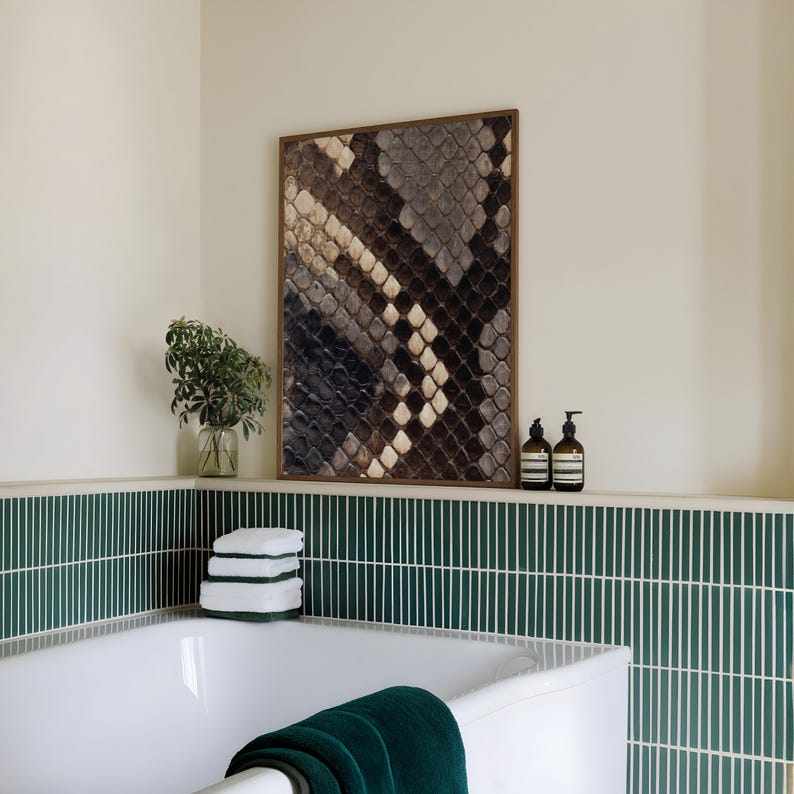 Op de afbeelding: Een rechthoekig wandkunstwerk met een gedetailleerd slangenleerpatroon in bruin-, grijs- en cr&egrave;metinten, gevat in een dunne houten lijst. Het kunstwerk hangt boven een wit bad met groene verticale tegels aan de muur.
